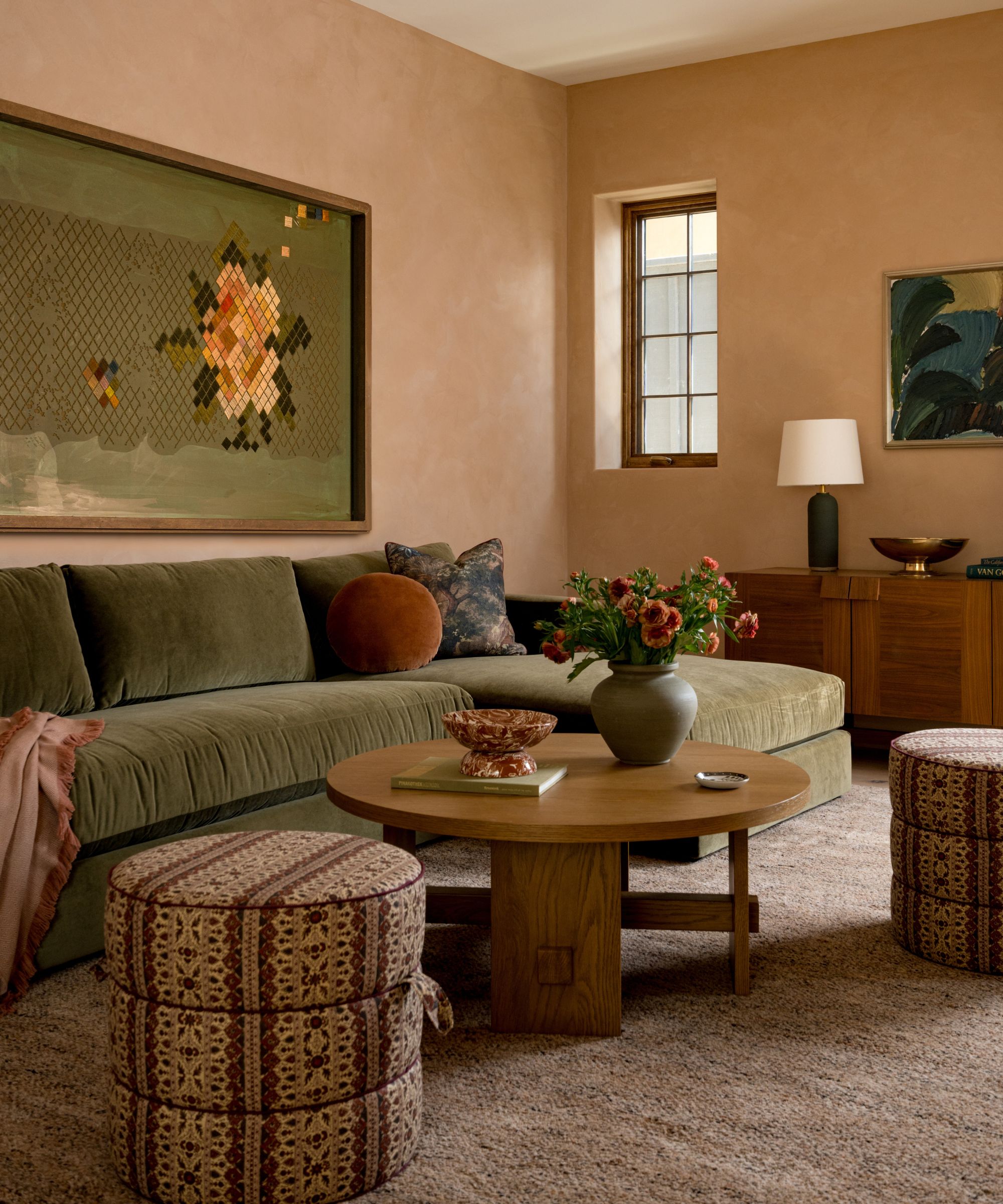 Warm living room with plaster pink walls, green velvet sofa, low wooden round coffee table, two floral ottomans, beige fluffy rug, and a large olive green painting of a yellow flower above the sofa