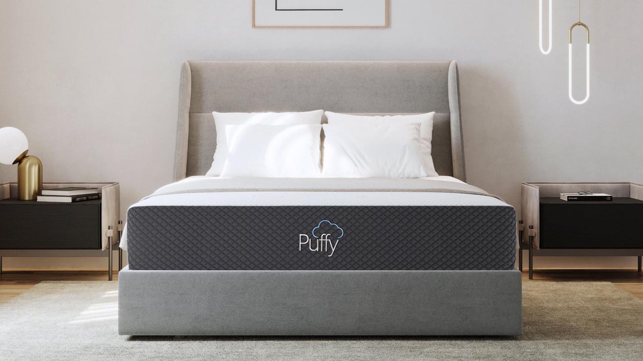 Best memory foam mattress: The Puffy Mattress with black base and white top, sat on a gray fabric bed frame and dressed with white bed pillows