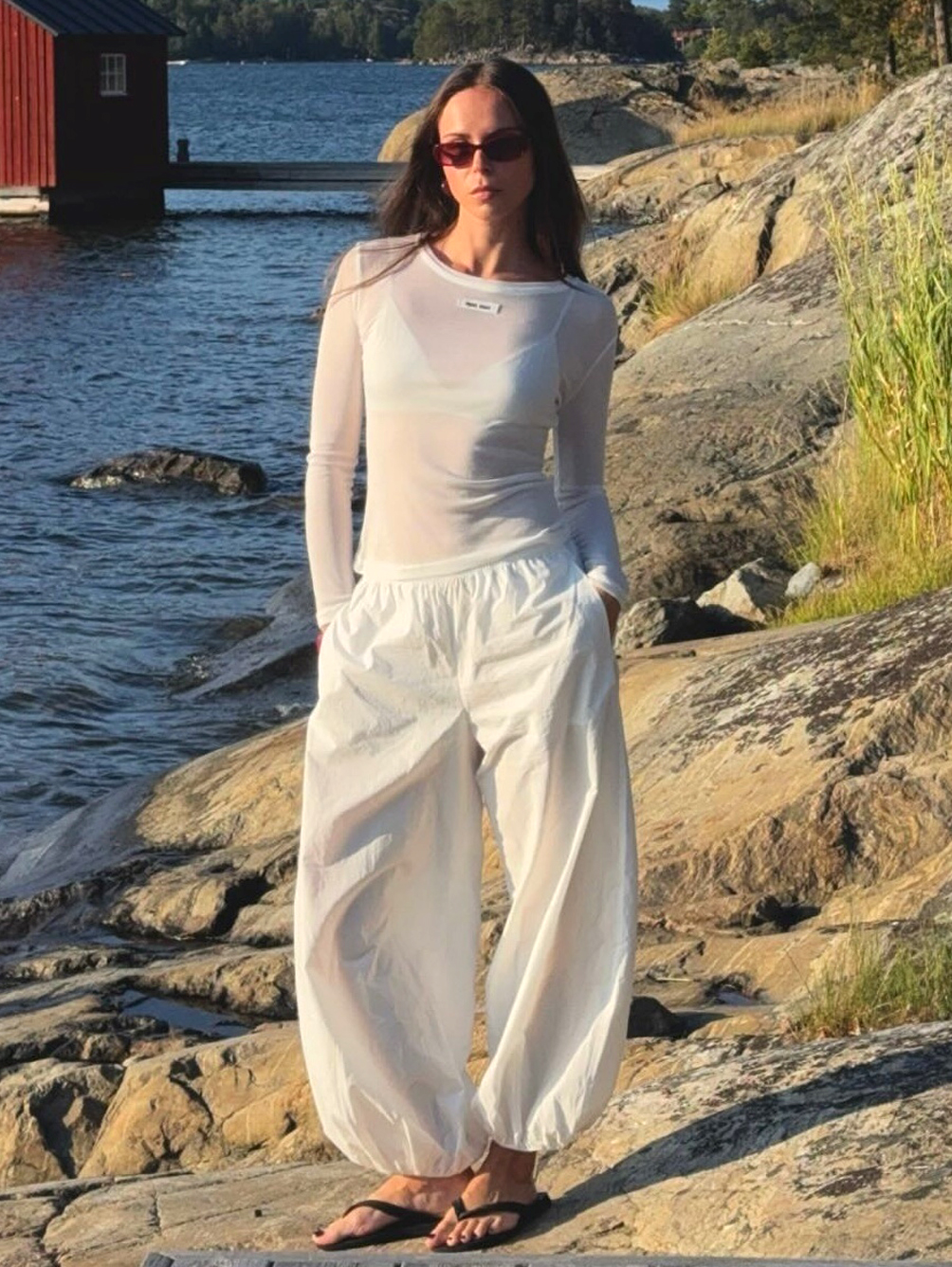 Swedish fashion creative Johanna Lager poses in Stavsn&amp;auml;s, Sweden on a rocky beach wearing red sunglasses, a sheer white long-sleeve T-shirt, white balloon pants, and black flip-flop sandals