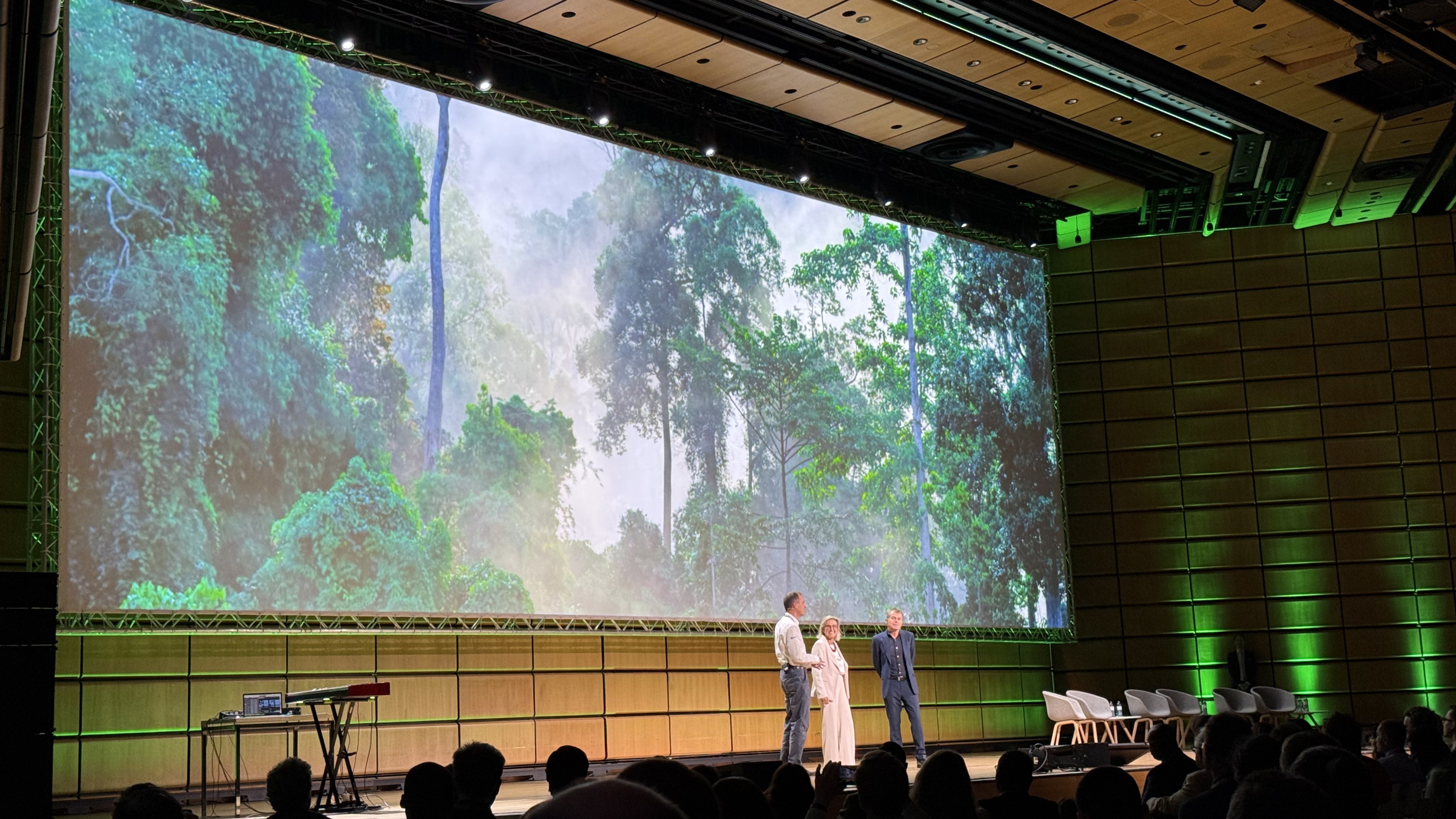 Three people stand on a stage in front of a screen with a forest scene on it