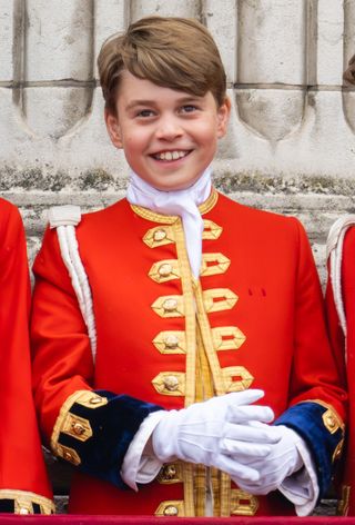 Prince George wearing a red and gold uniform with white gloves smiling while standing in front of a stone pillar