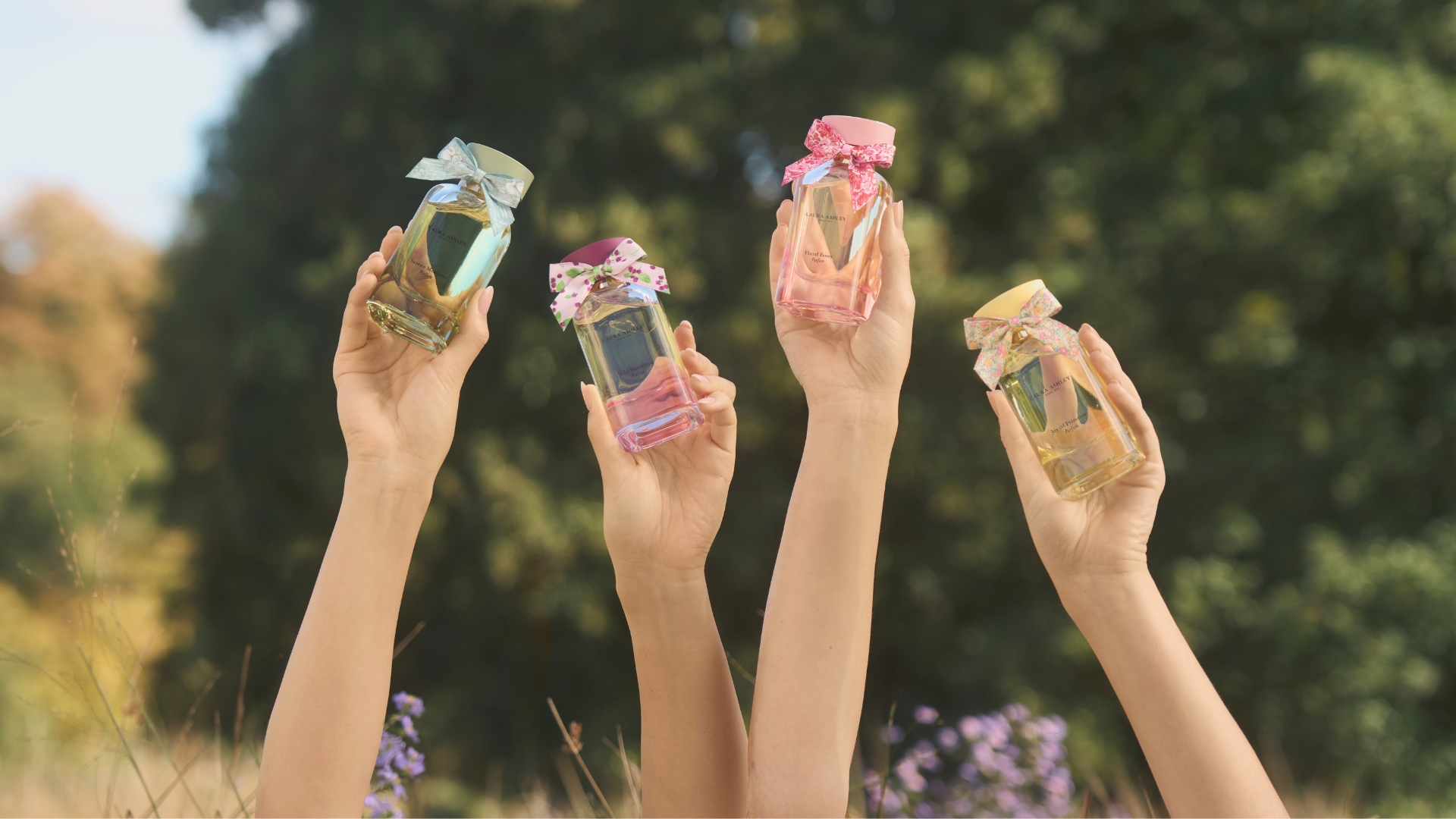Hands holding up the four bottles of new Laura Ashley perfumes with a field background