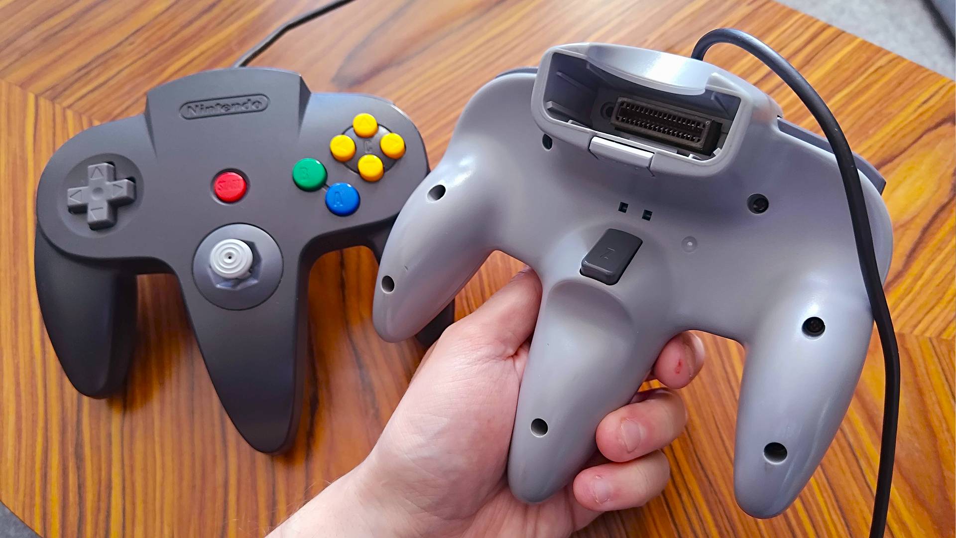 Hand holding gray N64 controller with Z button facing forward and black N64 gamepad on table in backdrop.
