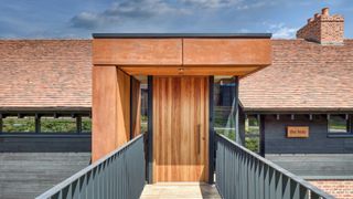 large modern iroko wood front door with corten steel porch on single storey long barn conversion with tiled roof and black weatherboard exterior