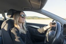 A young, blond woman in sunglasses drives a modern car with the sunset shining through her window.
