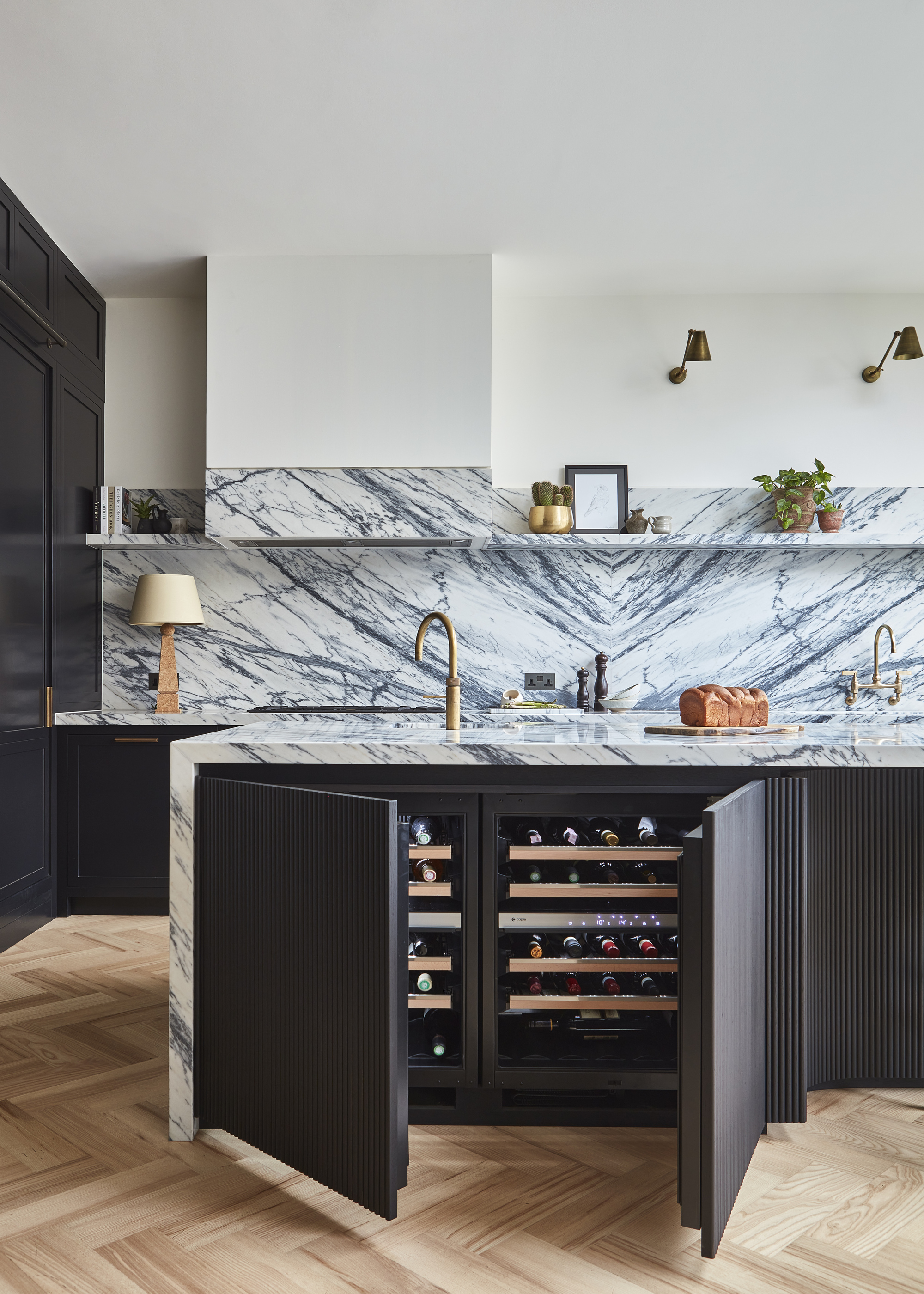 A kitchen with wooden herringbone flooring, black fluted cabinetry, and a book-matched marble countersplash. There doors of the kitchen island are open to reveal a wine fridge