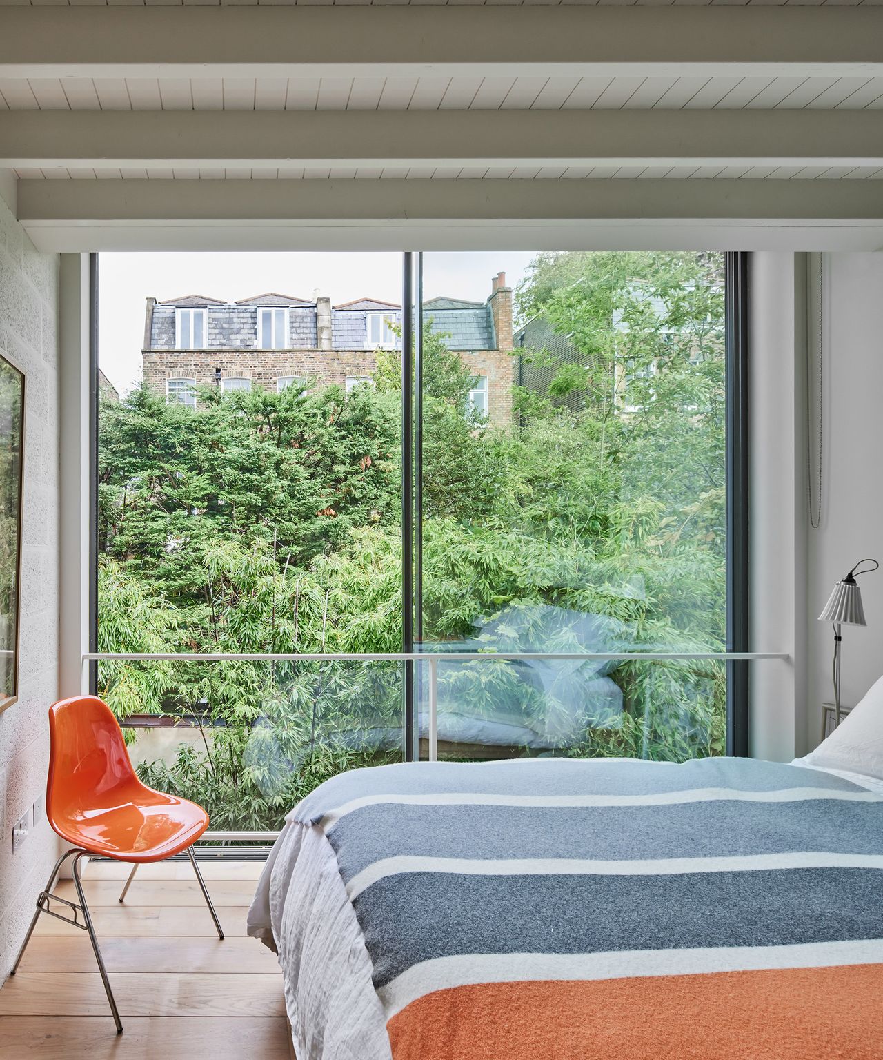 A small double bedroom with huge sliding floor-to-ceiling windows with colour block throw on the bed and bright orange chair