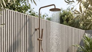 An outdoor shower in a brushed brass finish mounted against a pale slatted timber fence