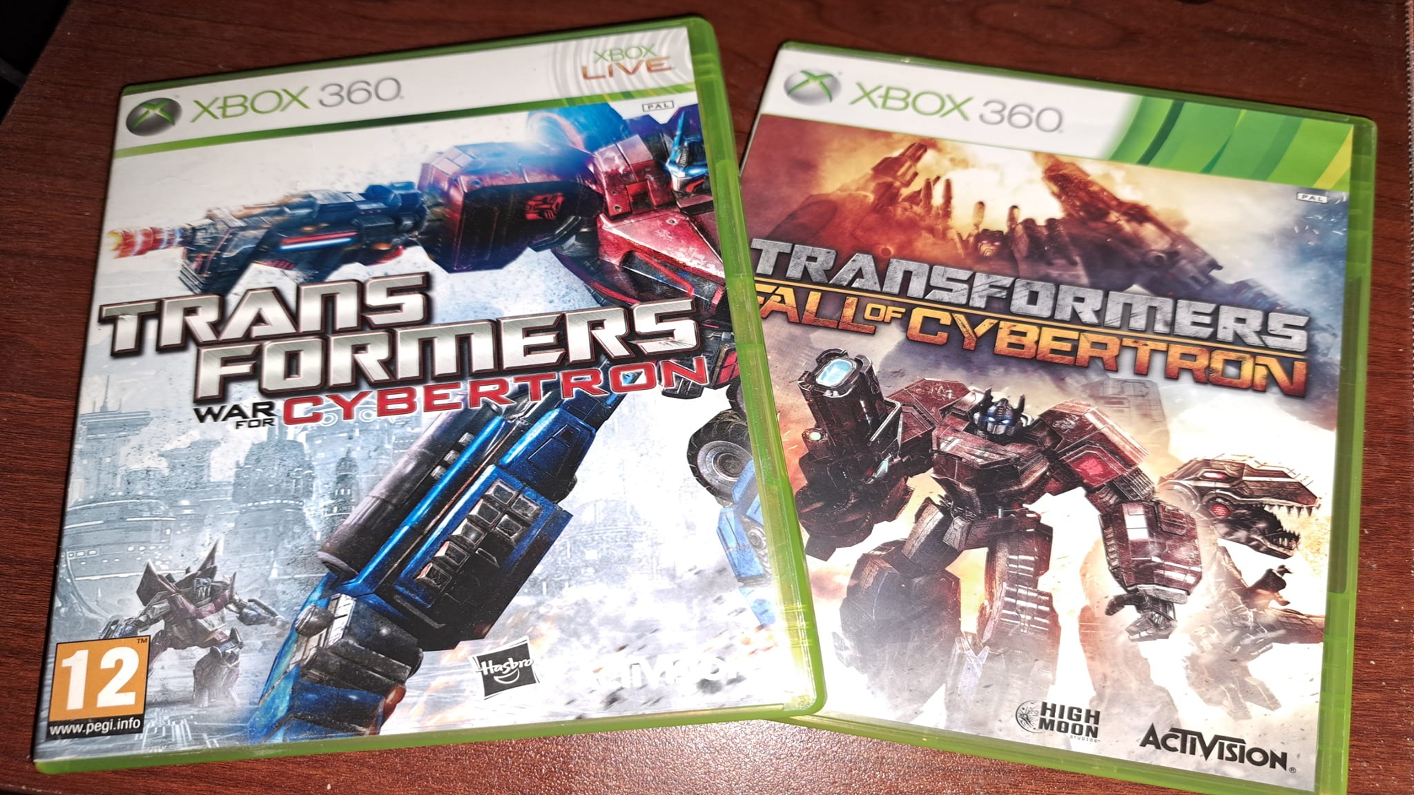 Two Xbox 360 game cases featuring Transformers: War for Cybertron and Transformers: Fall of Cybertron, displayed side-by-side on a wooden surface.
