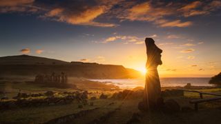 the sun sets behind a giant stone head on a lush green island