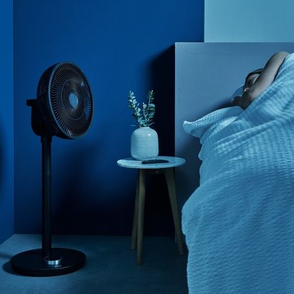 Duux Whisper Flex Smart Standing Fan on floor beside sleeping woman in blue bedroom