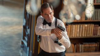 Michael Weatherly's Tony DiNozzo snazzily dressed and holding a gun while standing near a bookcase in NCIS: Tony & Ziva