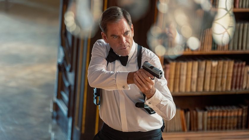 Michael Weatherly&#039;s Tony DiNozzo snazzily dressed and holding a gun while standing near a bookcase in NCIS: Tony &amp; Ziva