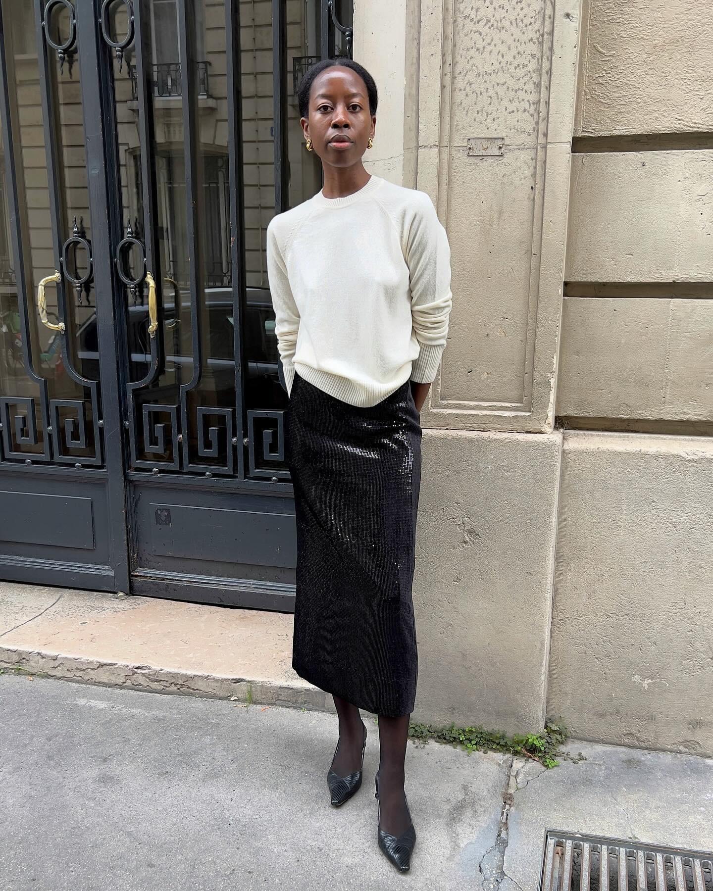 Influencer @sylviemus_ stands outside in Paris wearing a cream fine-knit jumper with a black sequinned pencil skirt and pointed-toe heels.