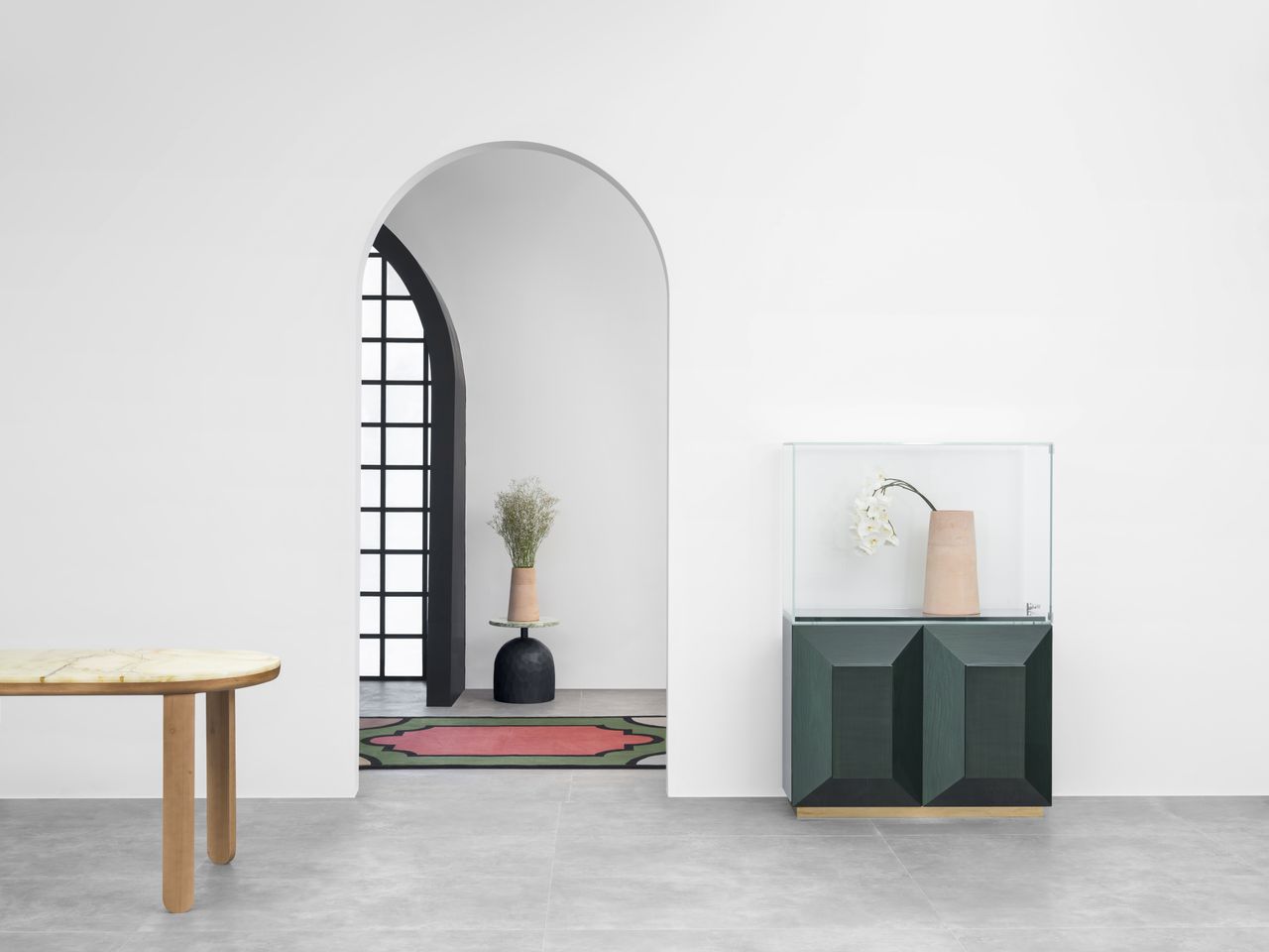 A pistachio table side, with a deep green cabinet with a glass container on top which has a vase with a flower in it, arched door through which we see a pink and green rug.