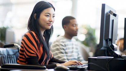 A young woman sits in front of a computer in a classroom with another adult student.