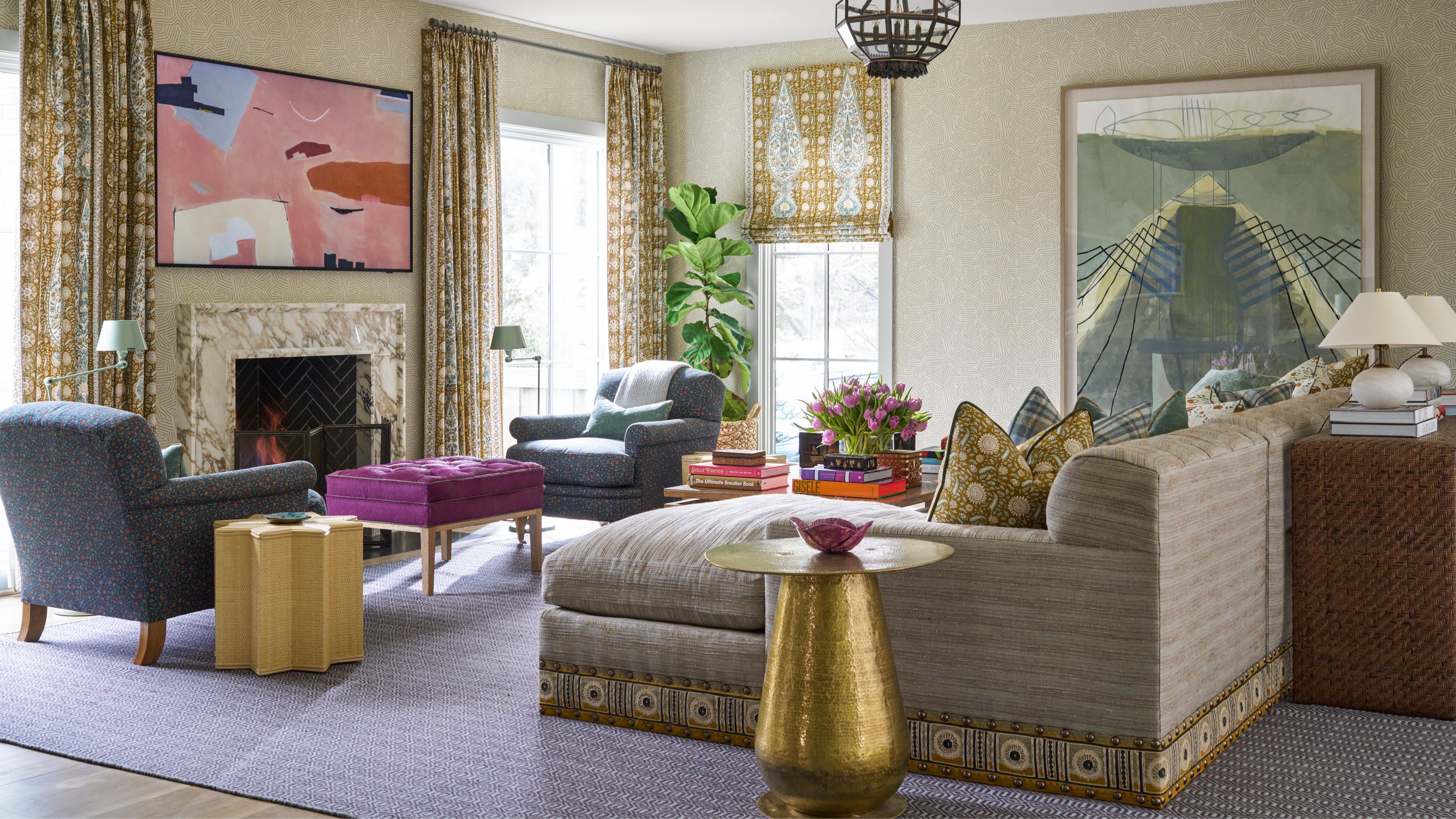 A bright and colorful living room centered on a long chaise sectional with neutral fabric and decorative embroidered trim. The room features a white marble fireplace, a vibrant pink abstract painting above the mantle, two blue armchairs, a rich purple ottoman, and large patterned curtains in yellow and gold hues. A large-scale abstract painting in green and blue hangs on the far wall.
