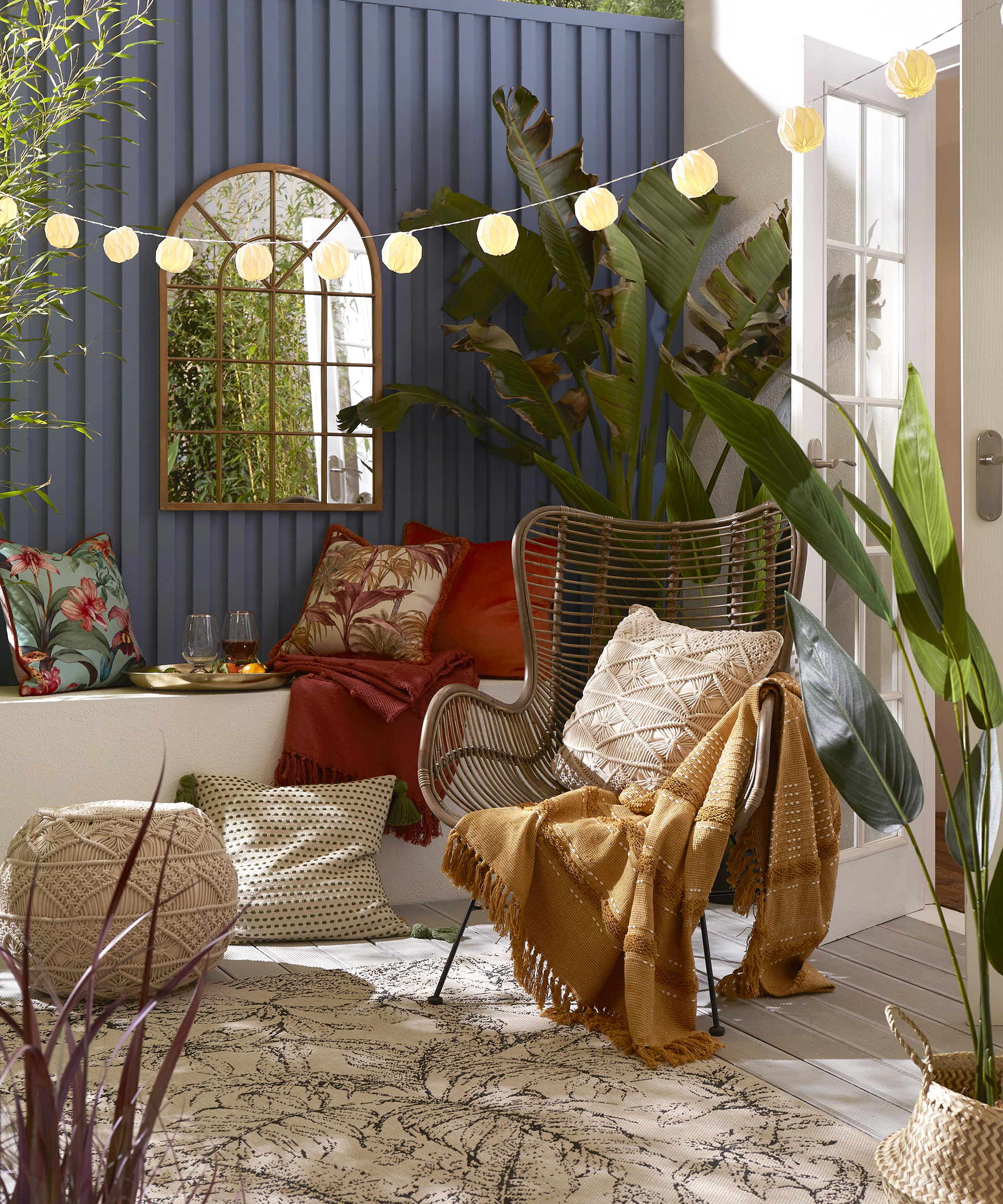 Boho garden with cream decking, LED festoon lighting, assorted colorful cushions, chair and houseplant with arched mirror by Joe Browns