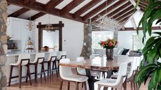 Wood kitchen ideas are so chic. Here is a kitchen with white walls and ceiling, dark wooden beams on the ceiling, white kitchen island and cabinets, and a wooden dining table with white seats underneath