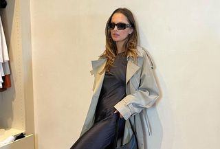Pia Mance leans against wall while wearing black slip and trench coat
