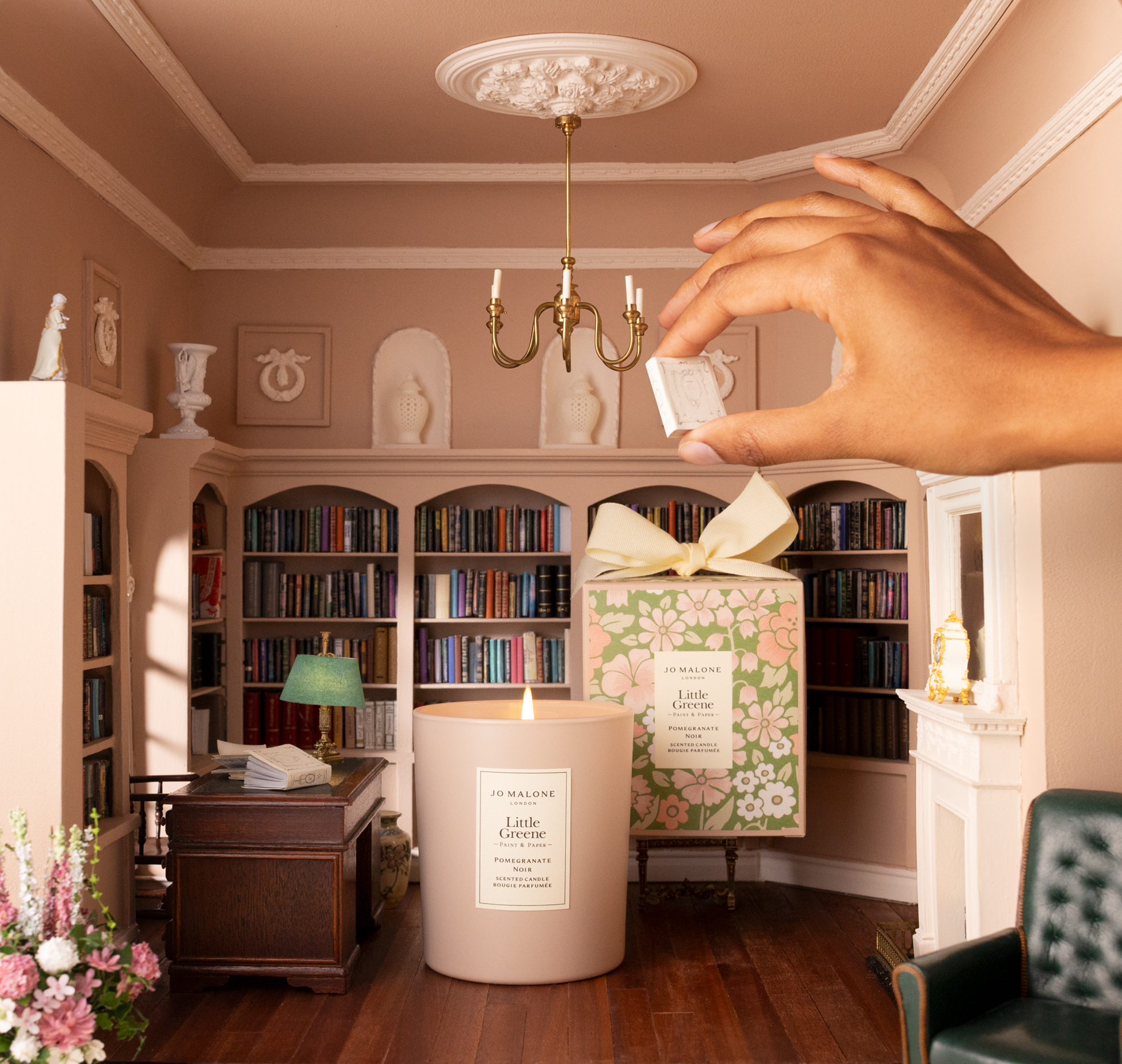 Image of a miniature room that is painted in a blush pink color with built-in bookshelves. There is a hand reaching in from the side to light the regular-sized candle that's in the middle of the room.