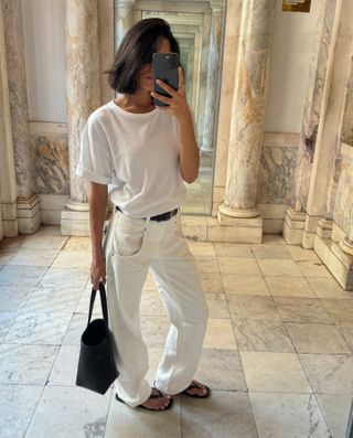 Shay wears a white tee, white jeans, black sandals, and a black bag.