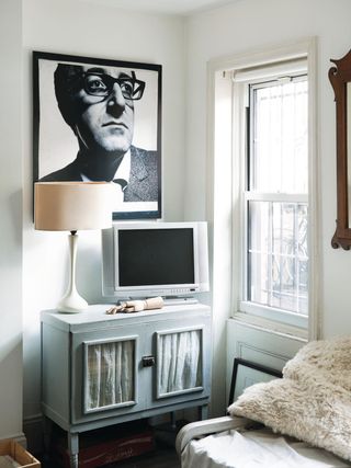 White living room with a blue cabinet that has fabric window covering inside. There is a black and white photograph hanging above the cabinet and a white lamp.