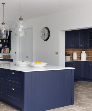 A kitchen with a dark blue kitchen island and white surface with two white bowls, white walls, and a door frame looking to dark blue cabinets and a brick wall