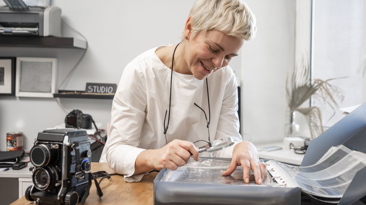 A woman uses a magnifying glass to look closely at photos in her photo studio.