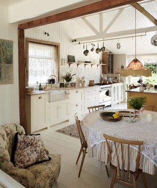 A white kitchen with off-white shaker cabinetry and shiplap wall, with wooden beams and round tableclothed table