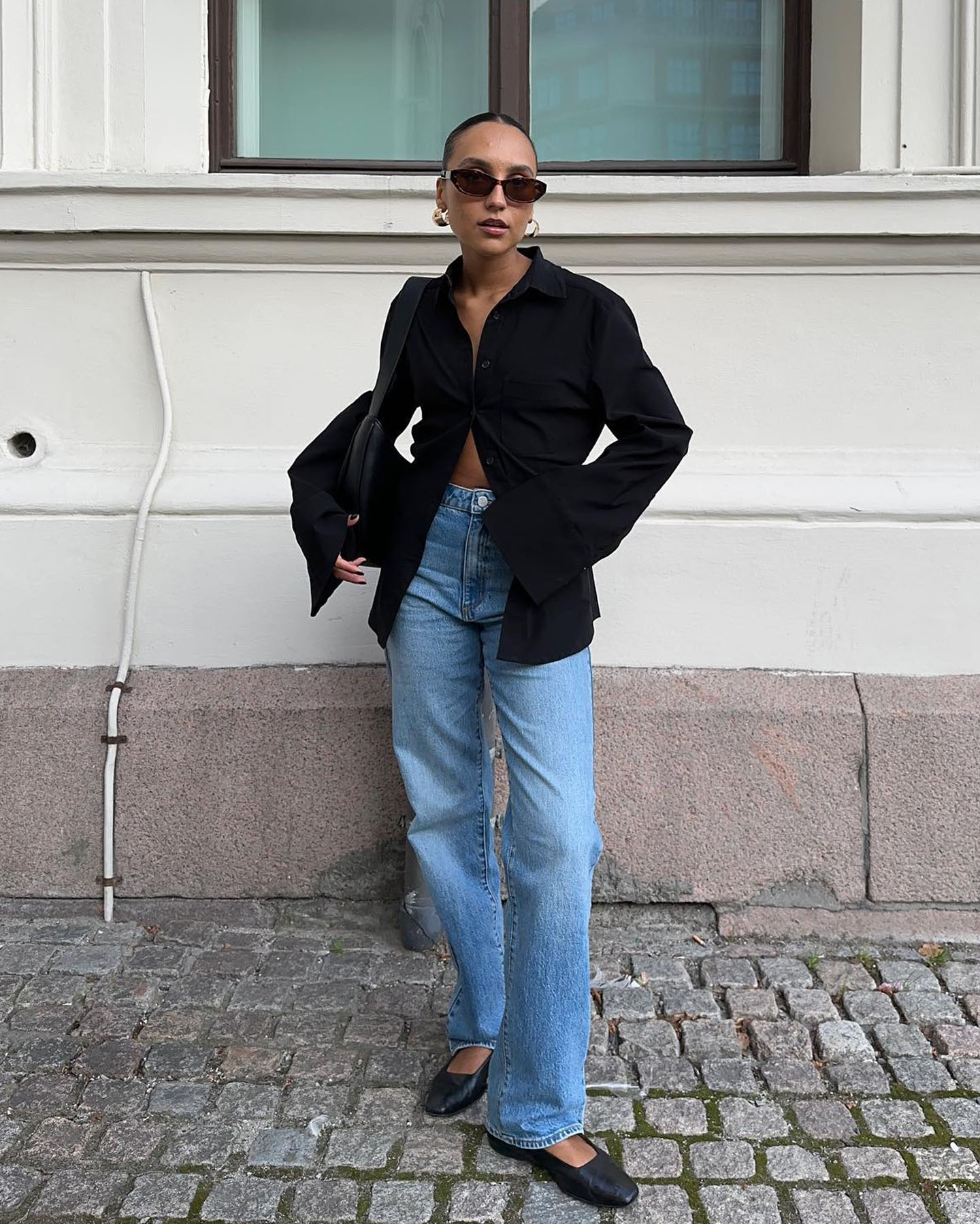 fashion influencer Ingrid Edvinsen poses on the street wearing hoop earrings, oval sunglasses, a black button-down shirt, jeans, and ballet flats