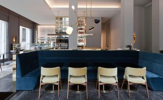 View of the interior of the restaurant with deep velvet blue and beige sitting area, a bar, gray and white marble flooring, and high windows.