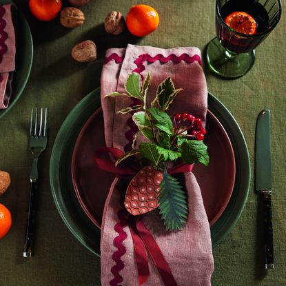 A dining table set for Christmas dinner with a green tablecloth and a pink wavy-edged napkin placed on top of plates