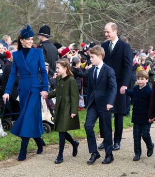 Kate Middleton walking outside past a crowd wearing a blue coat and holding hands with Princess Charlotte, who is wearing a green coat and walking next to Prince George, Prince William and Prince Louis