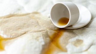 Image shows black coffee spilling out from a white cup onto a white mattress