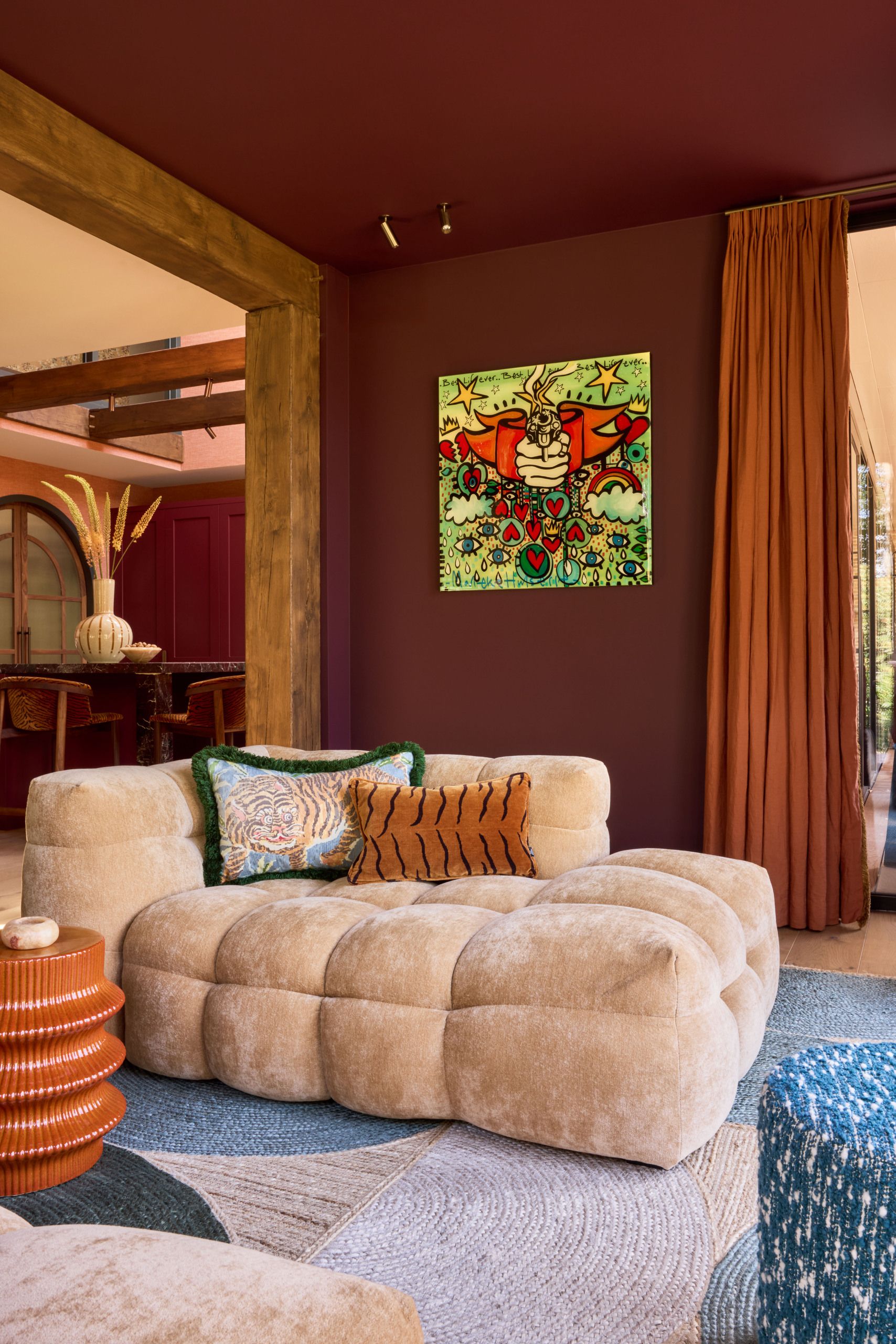 Image of a burgundy living room with a blue rug, orange curtains, an orange side table, and a low, puffy, cream-colored lounge seat.