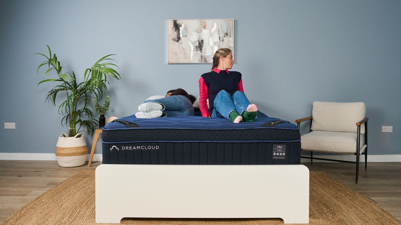 The image shows two mattress testers lying on a dark blue mattress, placed on a white bed frame and set within a muted blue bedroom