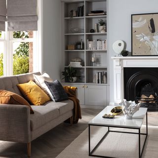 a living room with grey walls and grey inbuilt storage unit with shelves and cupboards, a bay window to the left and fireplace to the right, with a grey coach and a white top coffee table with black frame