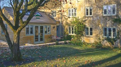 Exterior of extended Cotswold cottage on crisp bright autumn day. Fallen leaves and apples on lawn beneath apple tree.