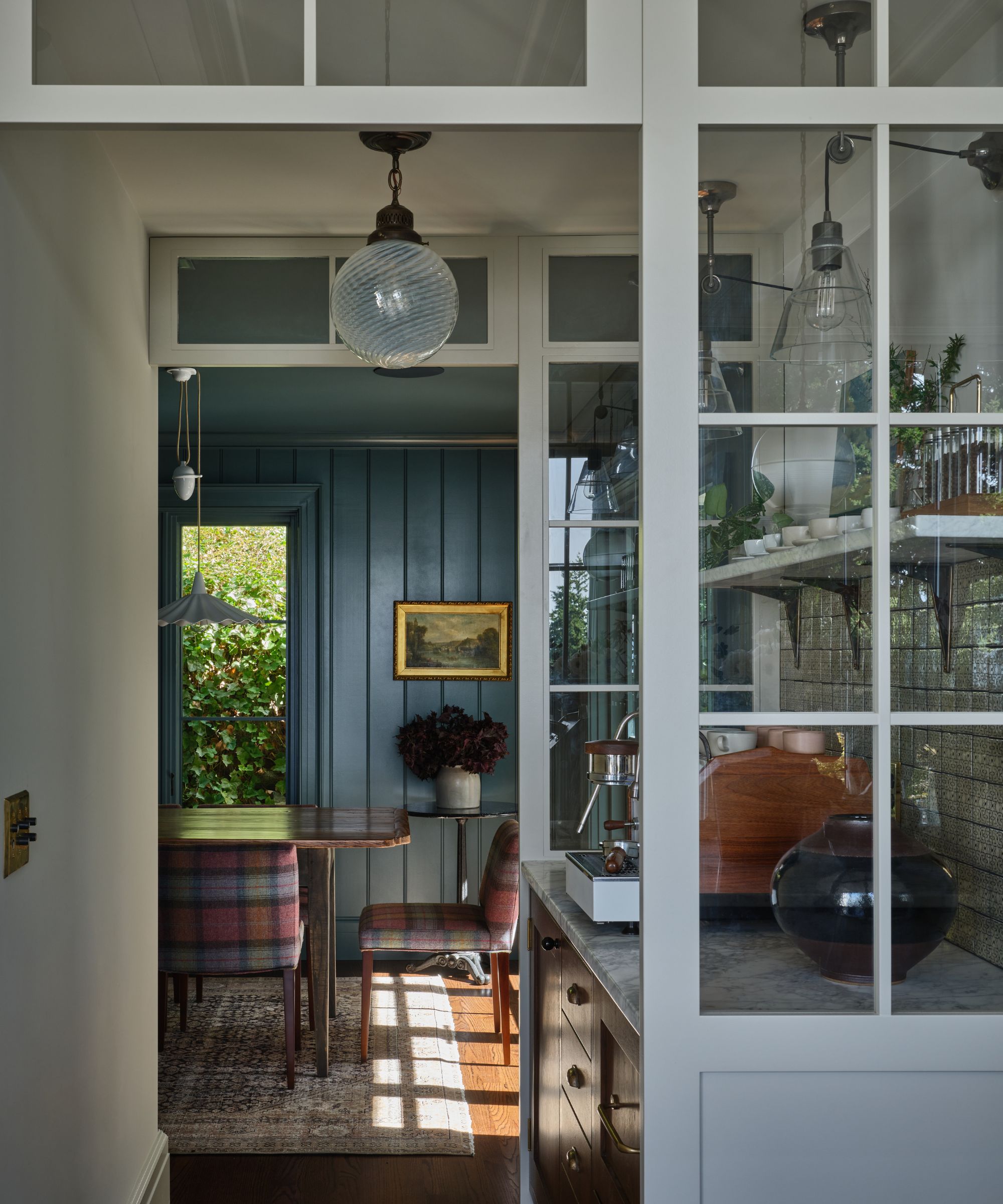 A looking into a moody teal dining room featuring plaid-patterned chairs and a rustic wooden table under a pendant light