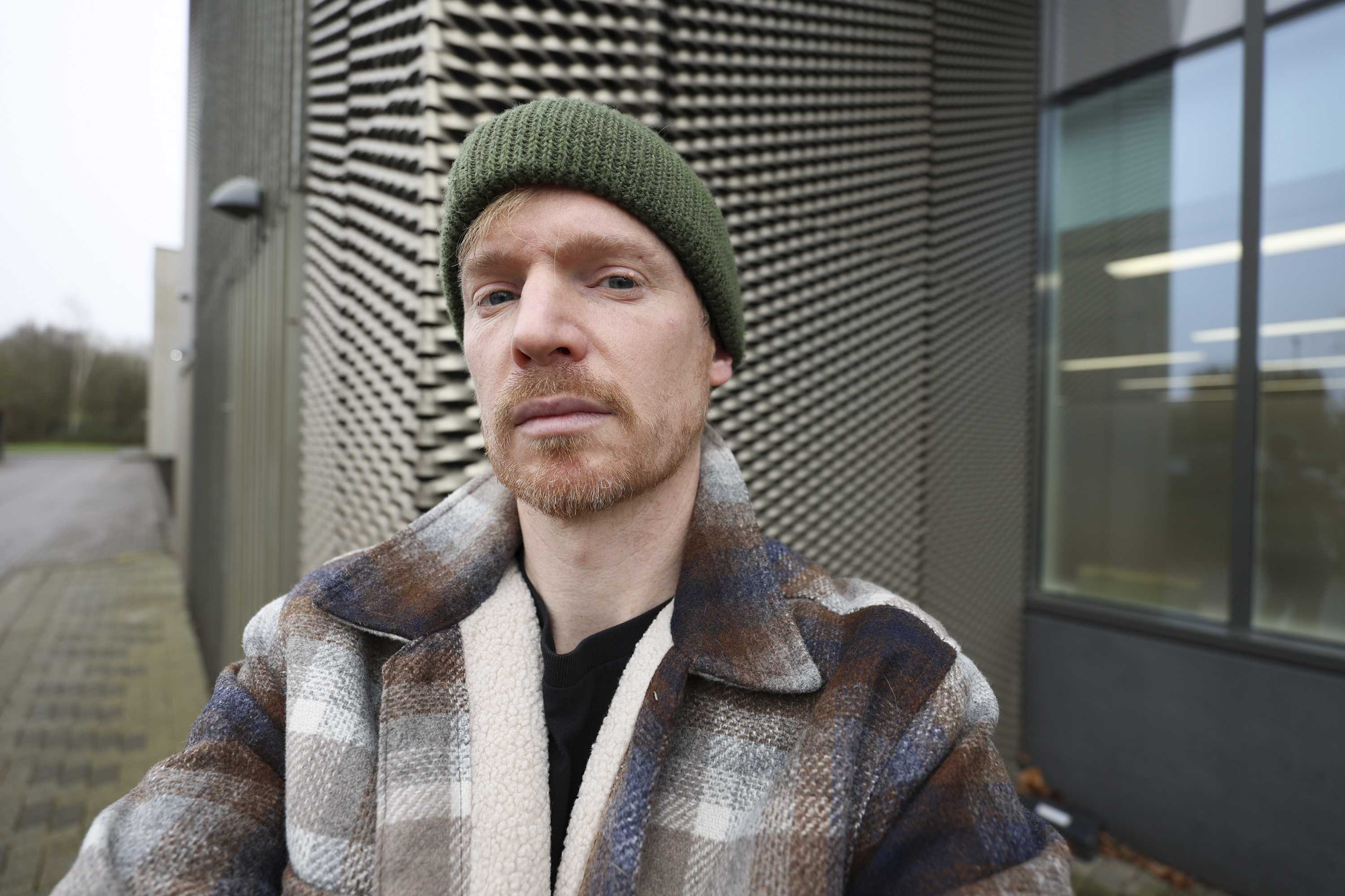 A man in a green beanie and shacket's selfie, outdoors in gray urban area