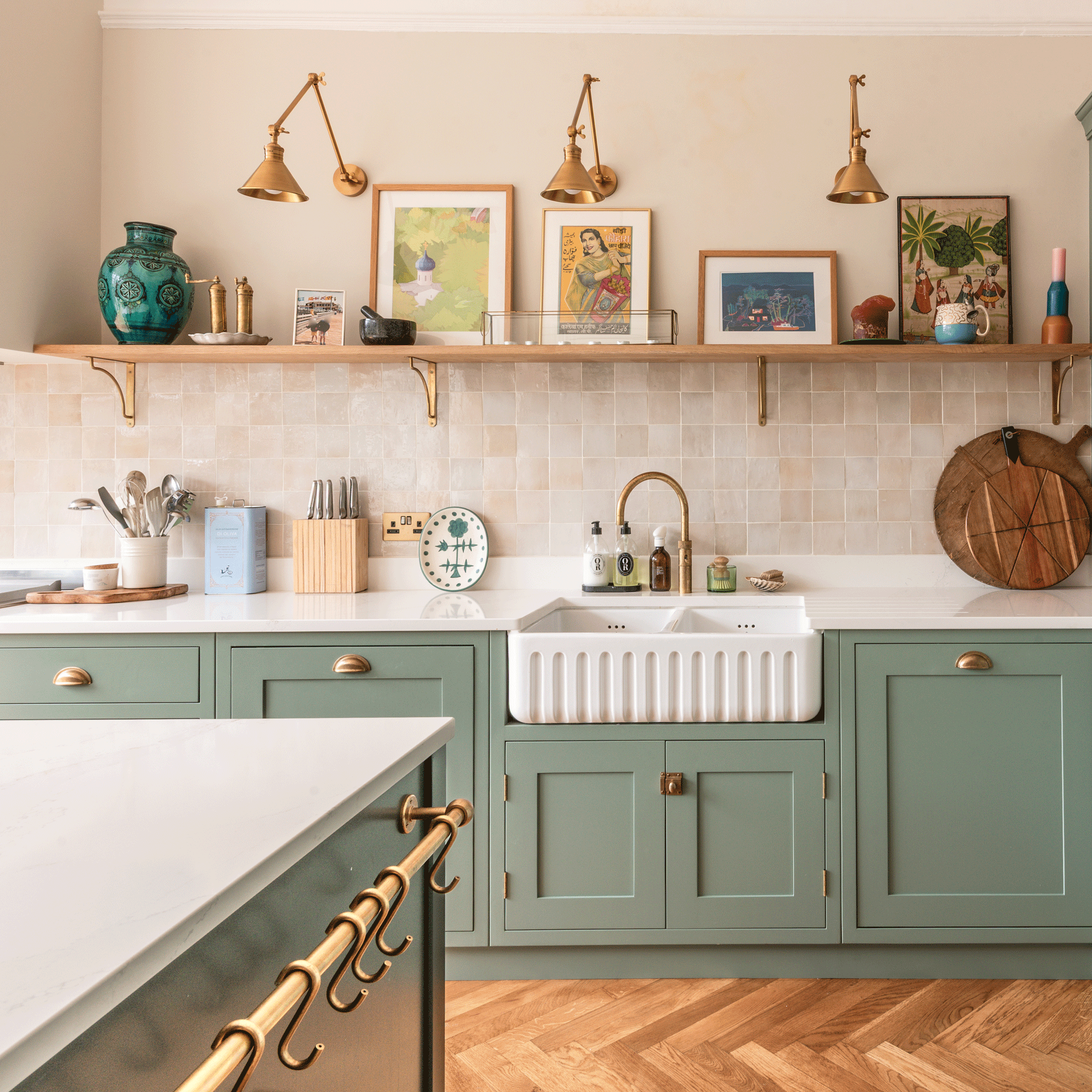 a kitchen with sage green cabinets and a fluted double Belfast sink with brass hot water tap, tiled splashback and open shelving, and a kitchen island with brass rail and s-hooks