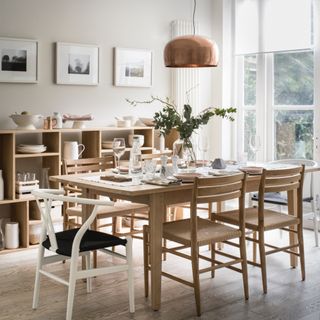 Scandi style dining room with blonde wood table and chairs with matching open plan storage unit with white tableware, copper pendant light above table, vase of eucalyptus, artwork, view of garden
