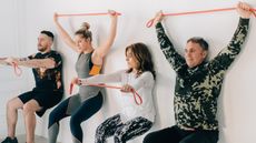 Four people exercising, in wall sit position holding orange resistance bands either in front of them or above their heads
