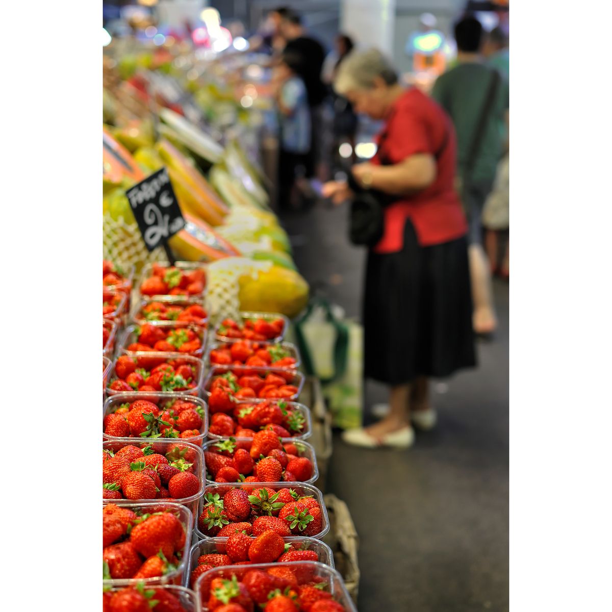 Tubs of strawberries in the foreground with blurred background showing person paying for items in Barcelona&rsquo;s historic La Boqueria market