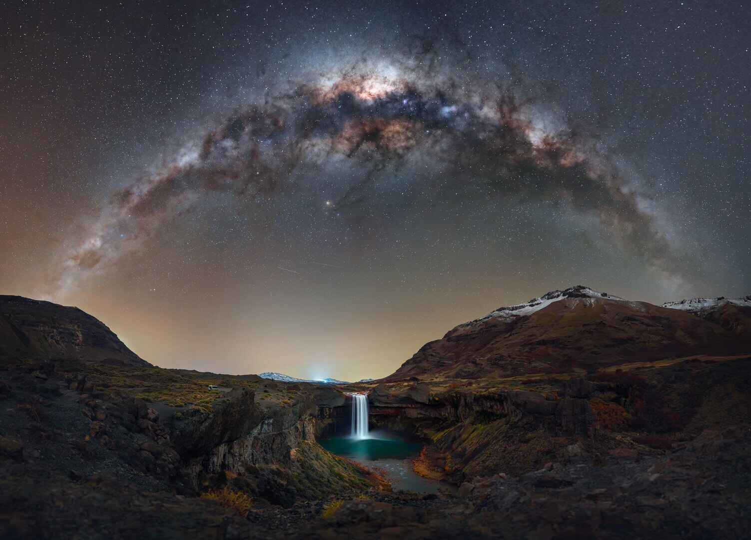 A waterfall is seen under the arc of the glowing Milky Way galaxy in the night sky.