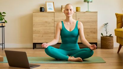 woman wearing a jade leggings and top set sat, eyes closed and crosslegged with hands in a yoga mudra on her knees, with a laptop in front of her in a living room setting. 