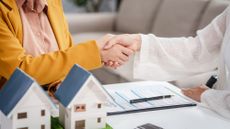 Two people shake hands over a desk with business contracts and two model houses