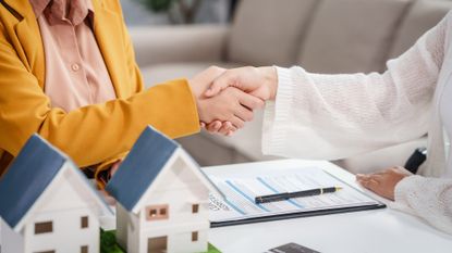 Two people shake hands over a desk with business contracts and two model houses