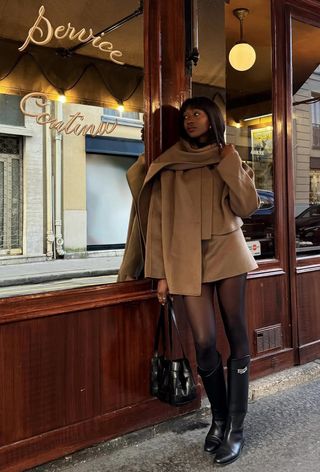 The winter capsule wardrobe item, knee-high boots, is shown in a photo of a woman standing against a restaurant storefront in pairs wearing a brown scarf coat with a matching mini skirt styled with black tights, black knee-high boots, and a black bag